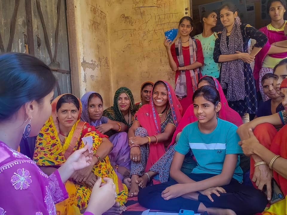 A group of women in saris sitting on the ground, creating awareness about period hygiene.