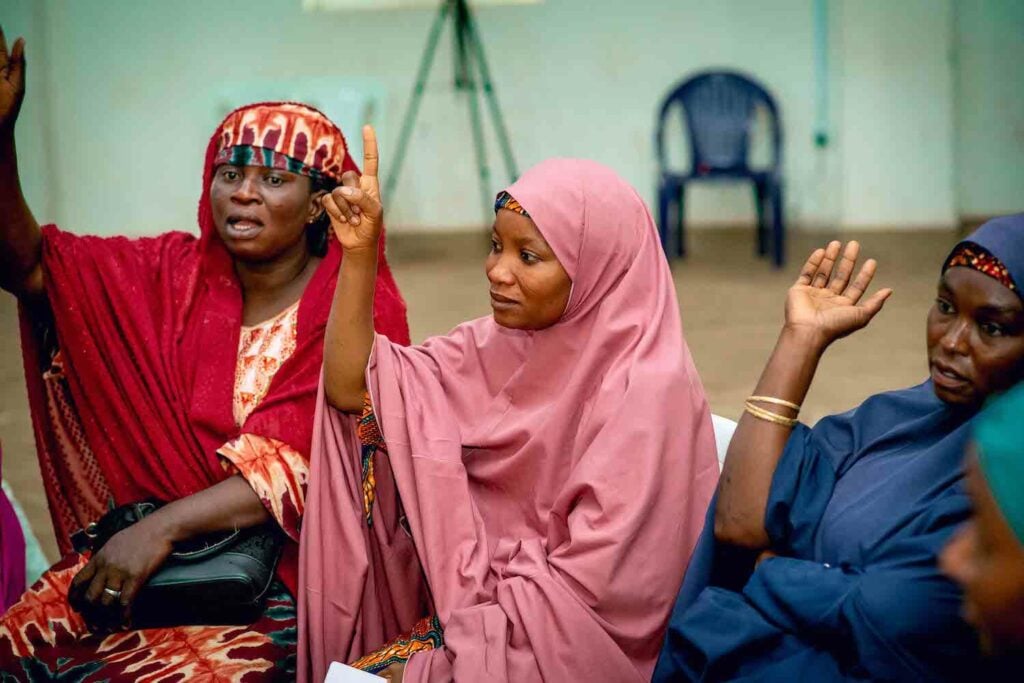 A group of women in hijabs are sitting and raising their hands, showcasing their participation in a Women's Cooperative Society for economic empowerment, supported by GPF Nigeria.