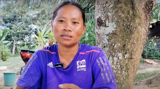 Woman speaking outdoors in a remote village with a tree in the background.