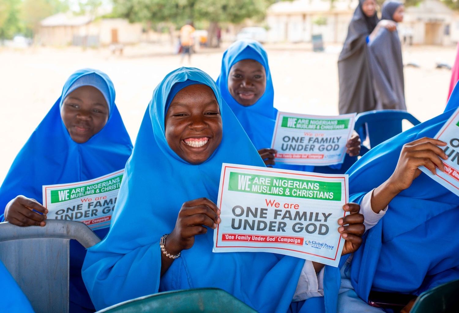 Group of smiling young students in blue hijabs holding signs promoting peacebuilding.
