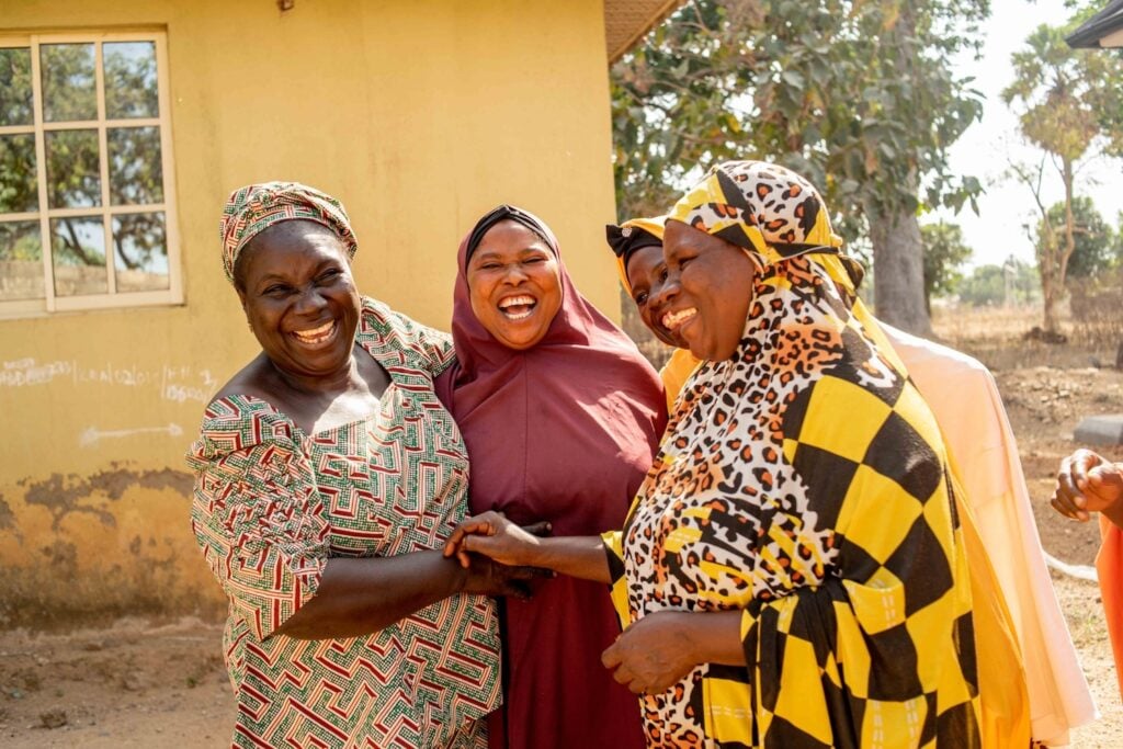 Four women, wearing colorful traditional attire, stand outside a building in Kagoro Chiefdom and laugh together while holding hands. Trees and a window are visible in the background, symbolizing the community resilience fostered through their interfaith dialogue.