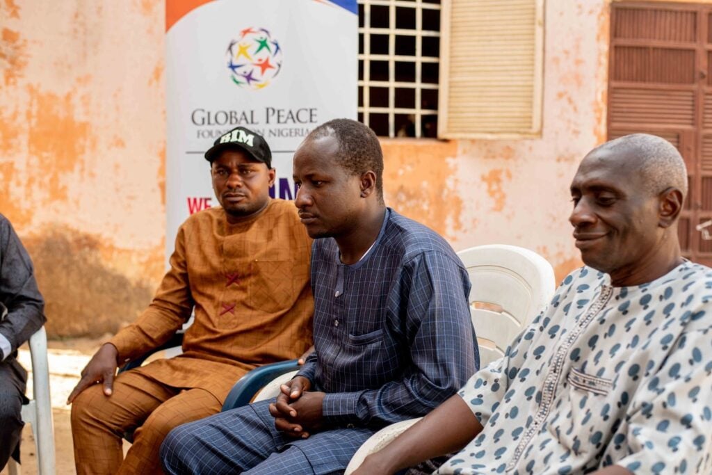 Three men sit in plastic chairs at an event by the Global Peace Foundation Nigeria; a banner is visible in the background, discussing Community Resilience and Interfaith Dialogue.