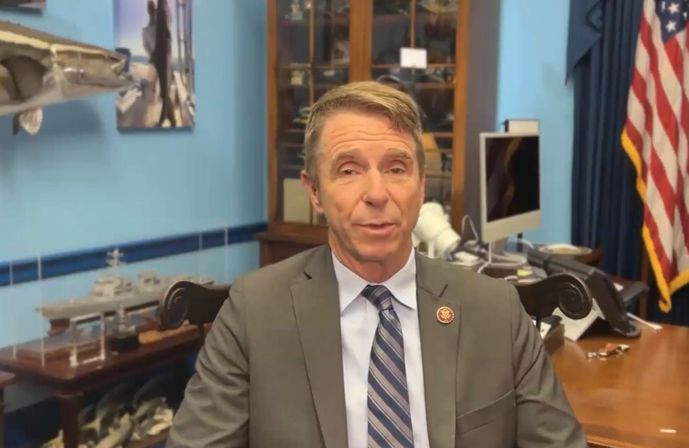 A man in a suit and tie is speaking in an office adorned with model ships, a computer, and an American flag in the background. He appears to be addressing the International Forum on One Korea.