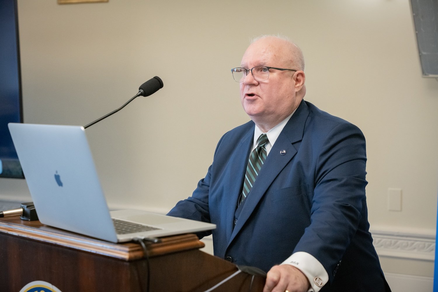 A bald man in a dark suit and striped tie speaks at a podium with a laptop, in front of a microphone, addressing the International Forum on One Korea 2024.