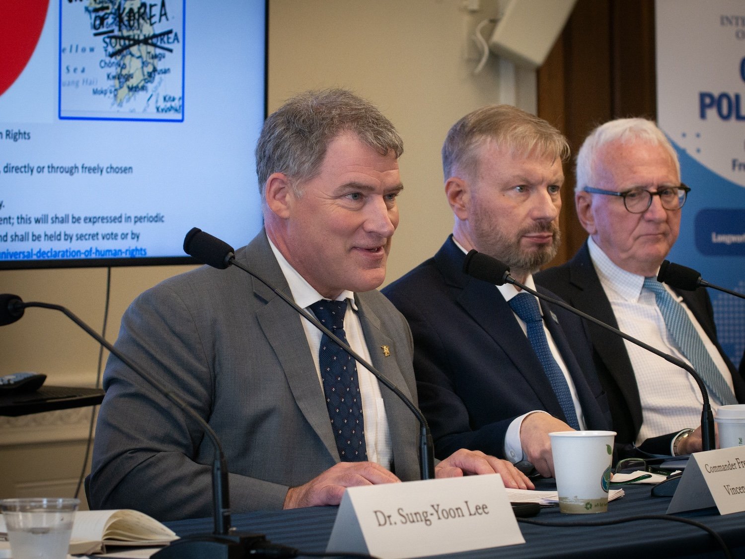 Three men in suits sit at a panel discussion with microphones in front of them, a screen with text and a map of South Korea in the background. A name tag reads