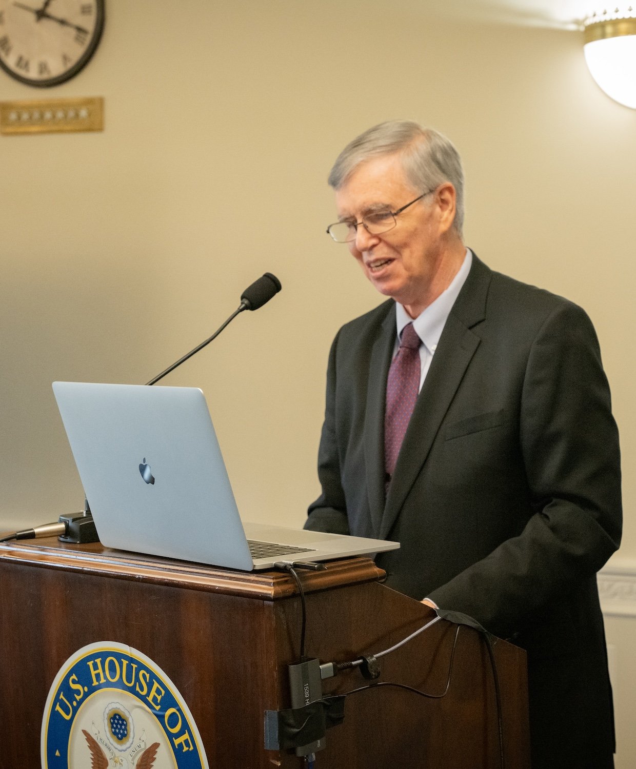 A man in a suit speaks at a podium with a U.S. House of Representatives seal, using a laptop and microphone during the International Forum on One Korea. A clock is on the wall behind him.