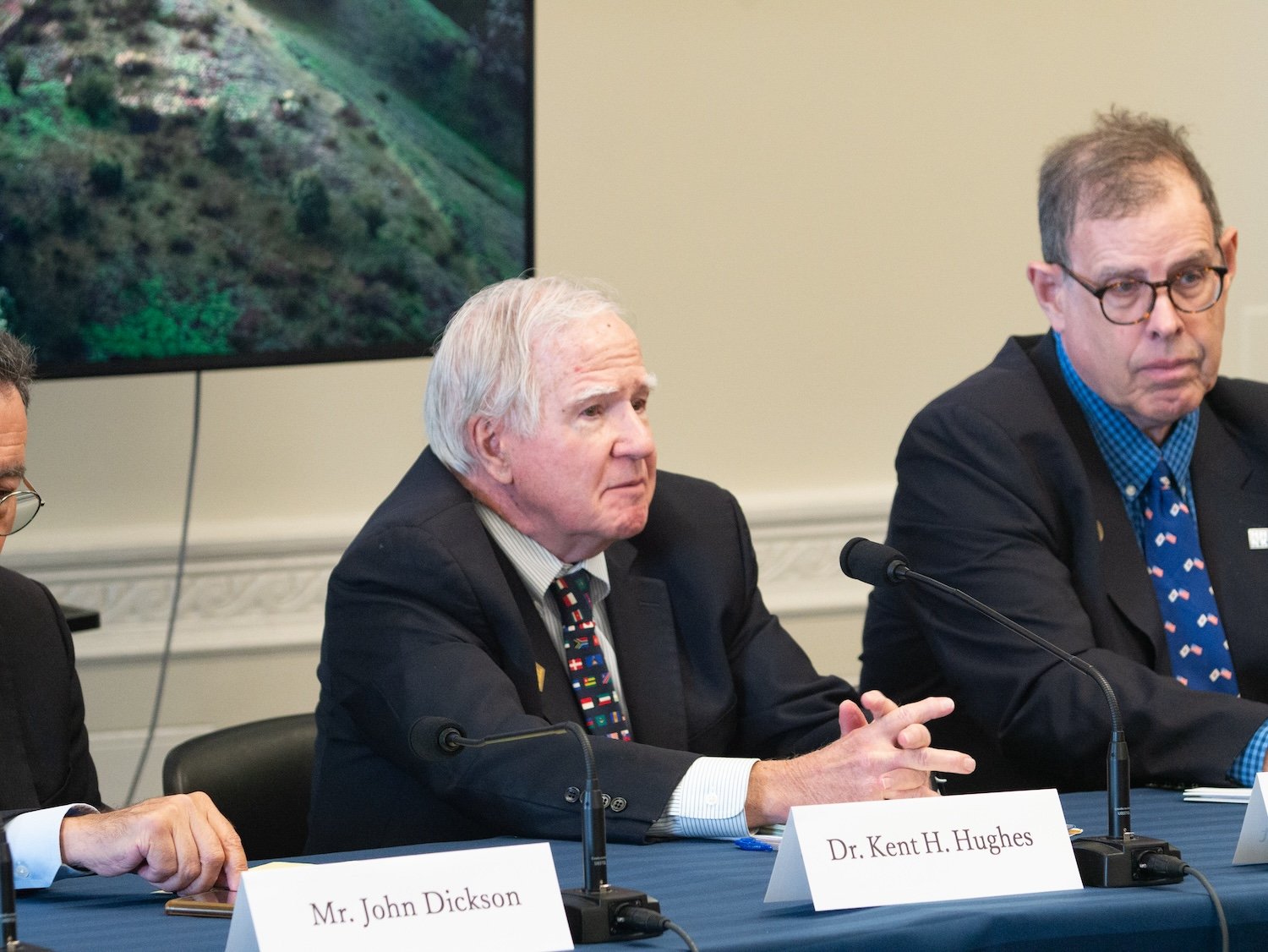 Three men in suits sit at a conference table with microphones; Dr. Kent H. Hughes and Mr. John Dickson have nameplates in front of them, participating in the International Forum on One Korea 2024 to discuss U.S. support for a Free and Unified Korea.
