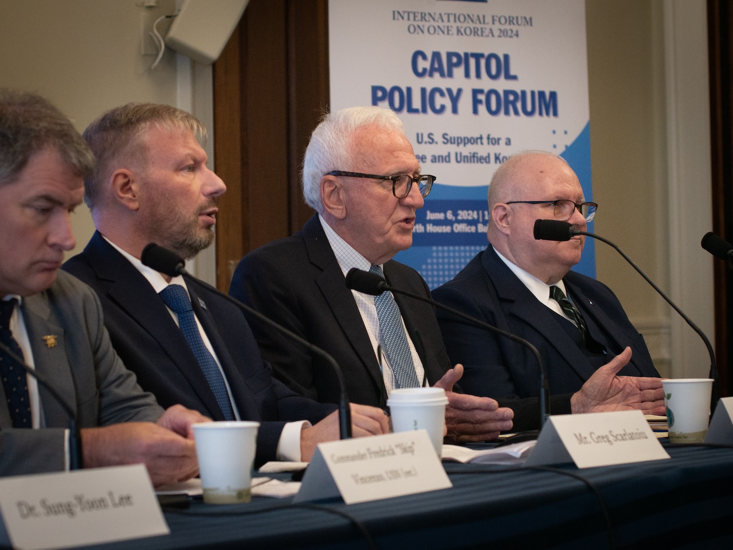 Four men in suits are seated at a table with microphones, papers, and coffee cups. Behind them is a banner for the