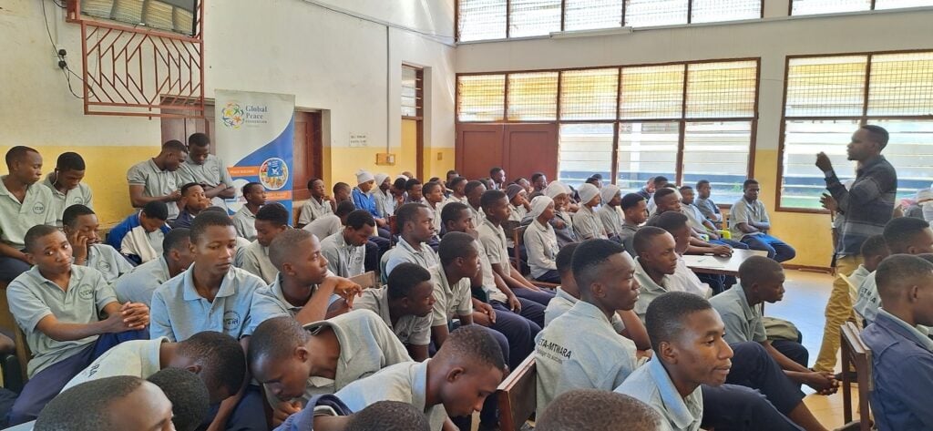 A person stands and speaks to a large group of uniformed Tanzanian youth seated in a classroom, delivering an engaging training session focused on business essentials.