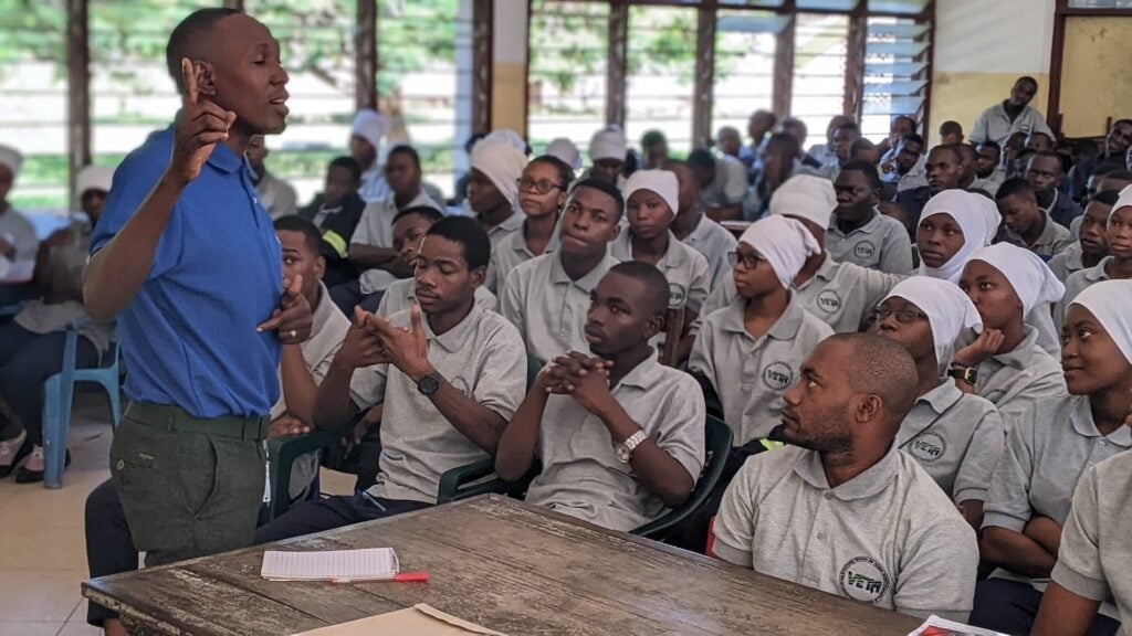 A person in a blue shirt speaks to a group of seated Tanzanian youth wearing gray shirts and white head coverings in a classroom setting. Papers and notebooks are on a desk in the foreground, suggesting an ongoing business training session.