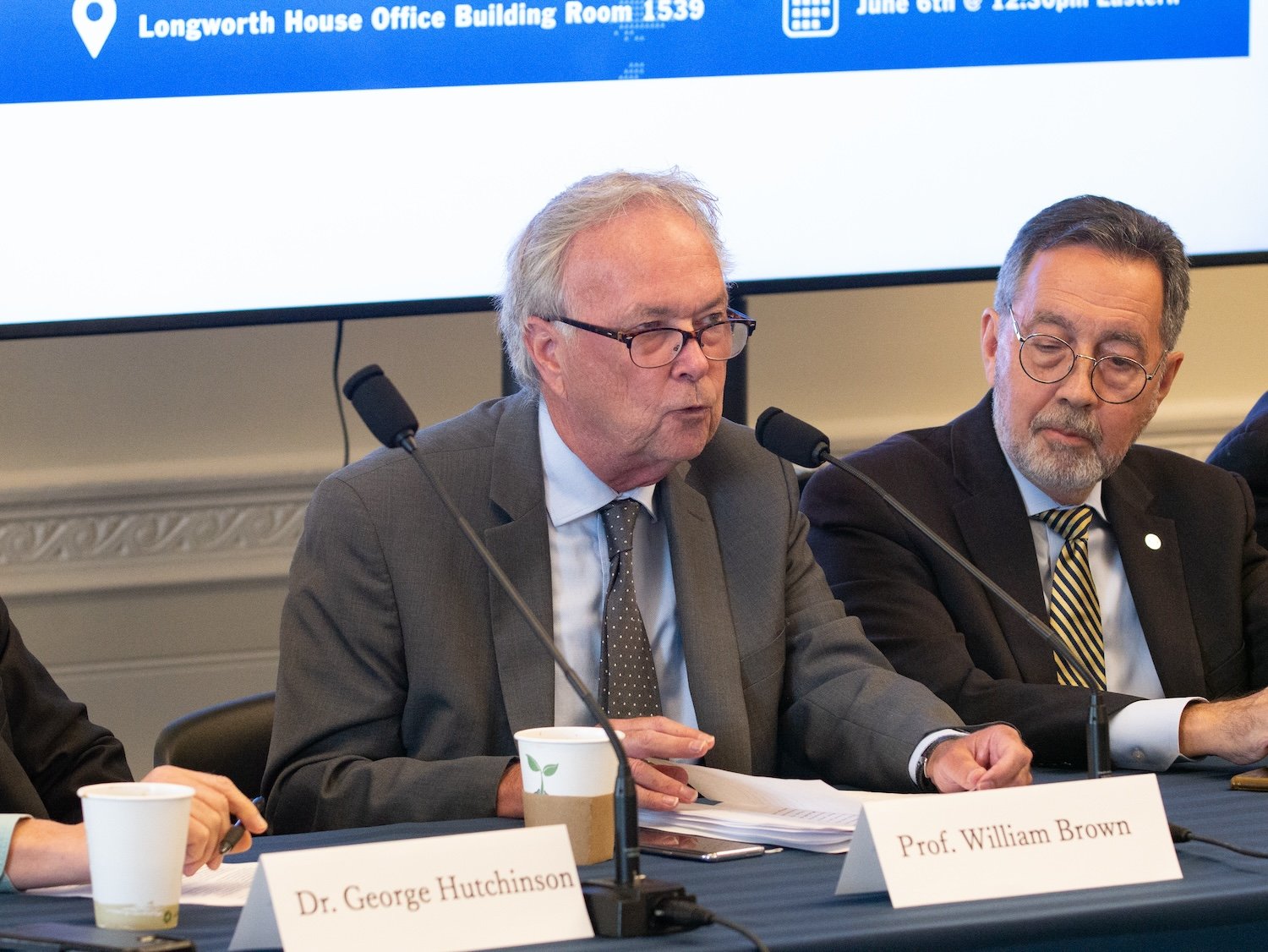 A man labeled Prof. William Brown speaks into a microphone at a conference table, discussing U.S. support for One Korea 2024, with another man beside him. A name tag reading Dr. George Hutchinson is visible on the table.
