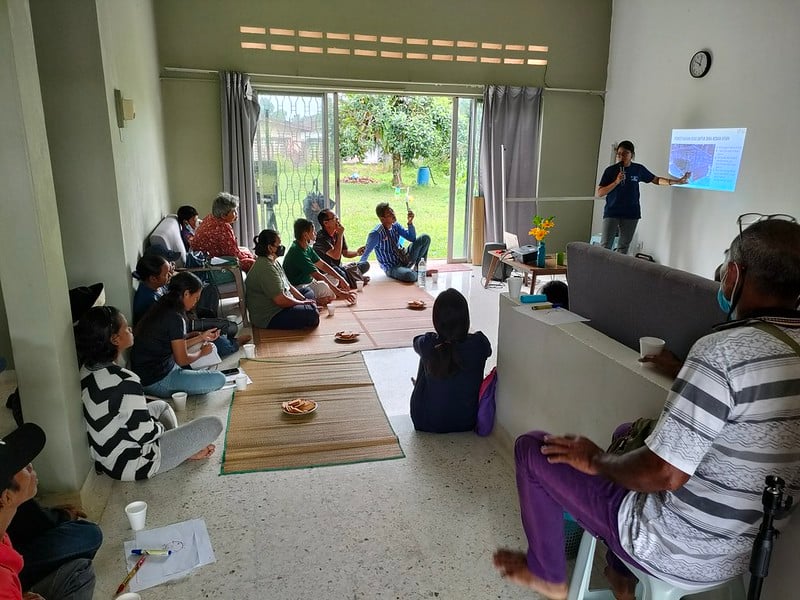 A group of people sits on mats and chairs in a living room while a person at the front gives a presentation with a projected slide on the wall, discussing sustainable chicken farming techniques and their impact on rural communities.