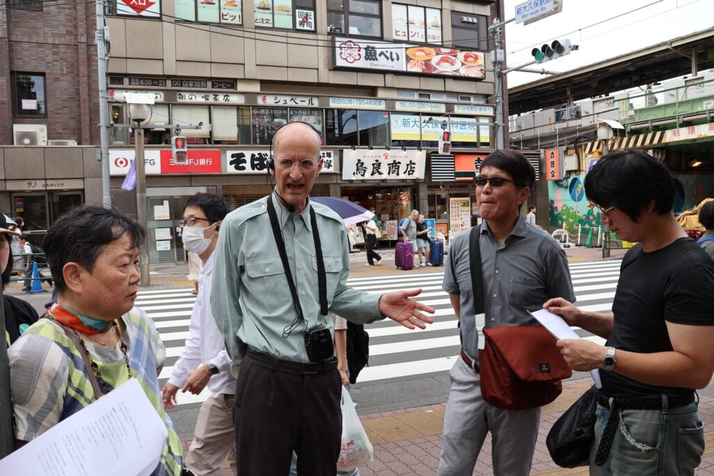 A group of people, some holding papers, stands on a sidewalk near a crosswalk in Shin Okubo, an urban area with shops and signs. One man, with a camera around his neck, appears to be speaking to the group likely about Asian history.