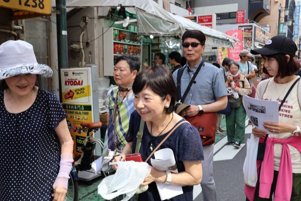 A group of people walking down a street in Shin Okubo, some holding maps and papers, in an urban setting with storefronts and signs in the background, reflecting the vibrant tapestry of Asian history.