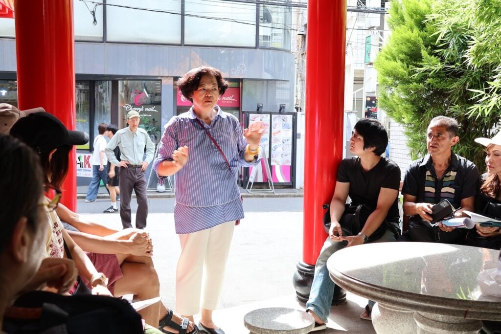 A person is speaking to a seated group outdoors near red columns. In the background, a busy street with pedestrians and a building with signs hint at the vibrant Shin Okubo area, rich in Asian history.