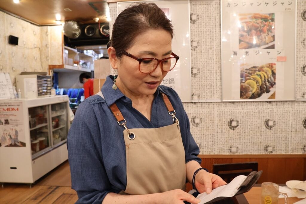 A woman wearing glasses and an apron is reading a menu or order pad inside a restaurant. The background includes a refrigerator, utensils, and images of Asian history on the wall. It’s reminiscent of the quaint eateries you’d find during a walk in Shin Okubo.