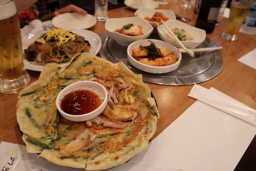 A table with Korean dishes, including pajeon (savory pancake) with dipping sauce, japchae (stir-fried glass noodles), kimchi, and other side dishes. Chopsticks and drinks are also visible. The setup evokes a sense of dining in Shin Okubo, rich in Asian history and culinary tradition.