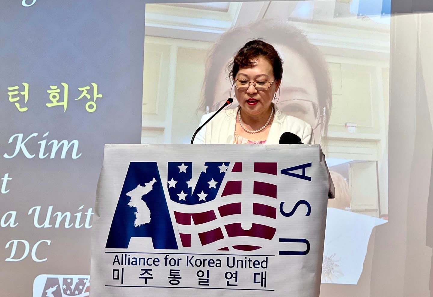 A woman stands at a podium speaking into a microphone. The podium features a banner reading