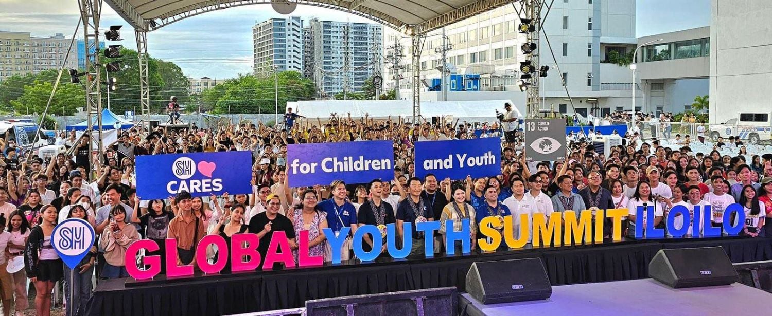 A large group of people stands on stage under a tent, holding signs that read