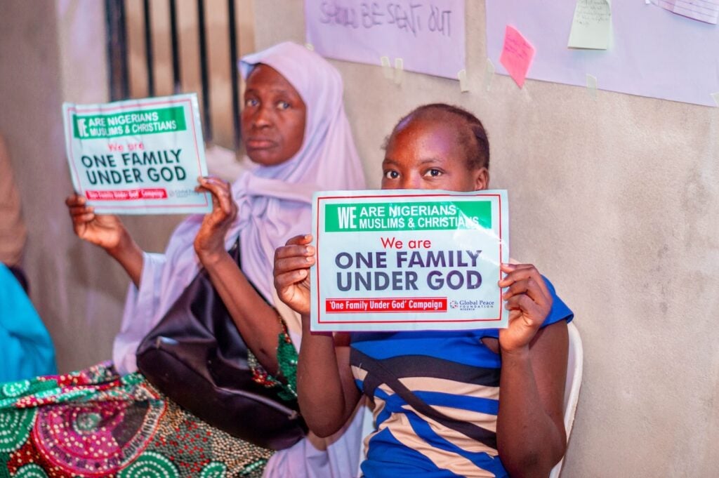 Two people sit, each holding a sign that reads