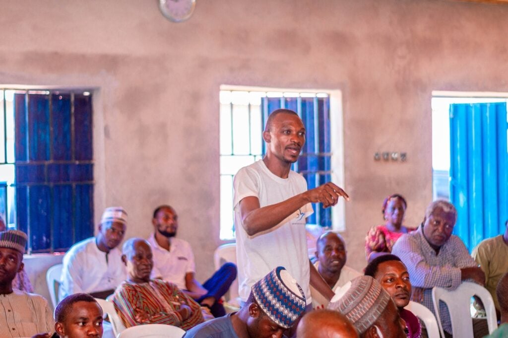 A man stands and speaks during an interfaith peacebuilding meeting in a room filled with seated attendees. The room has blue window coverings and a clock mounted on the wall.
