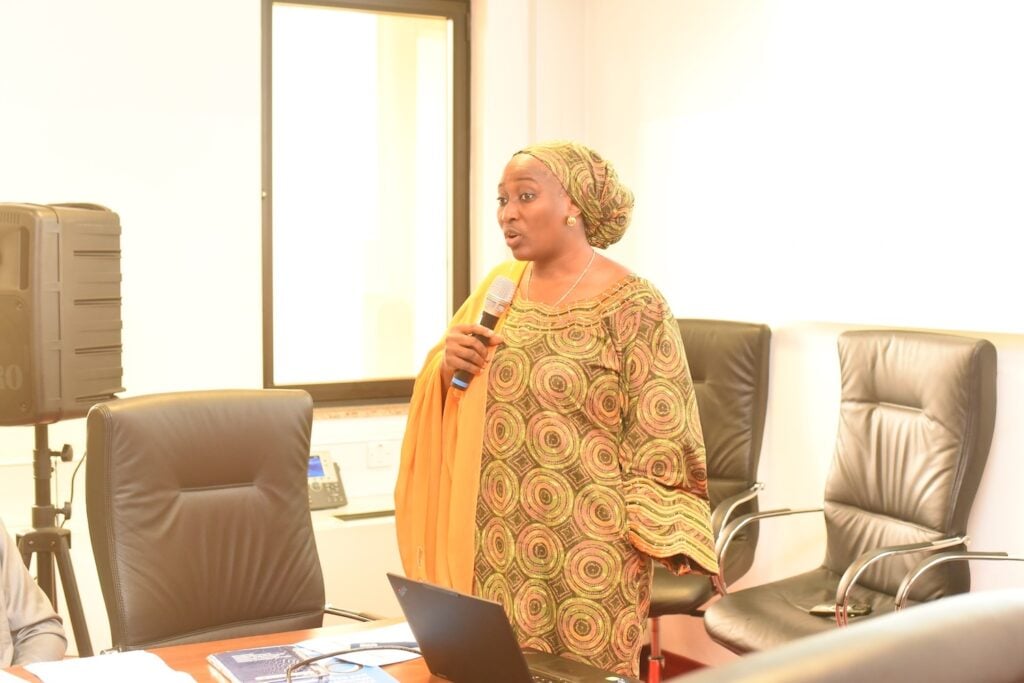 A person in patterned attire holds a microphone, speaking in an office with empty chairs and a laptop on a table, emphasizing the importance of interfaith dialogue for promoting religious tolerance.