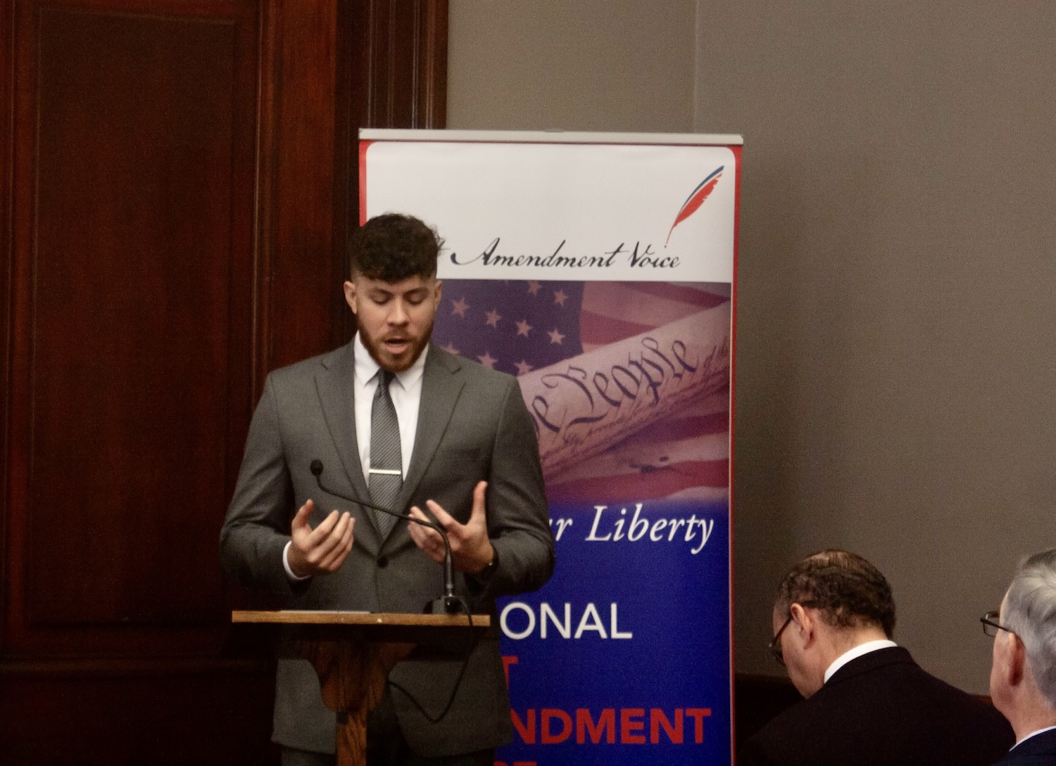 A man in a suit stands at a podium on Capitol Hill, addressing the importance of free speech against a backdrop featuring patriotic imagery and text. Several people are seated nearby, listening intently.