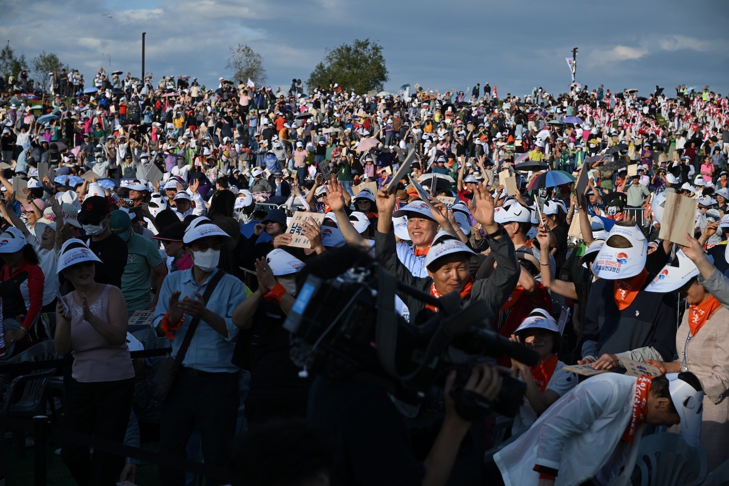 A large crowd of people, united under the Korean Dream, wears sun visors and hats, raising their arms and cheering outdoors.