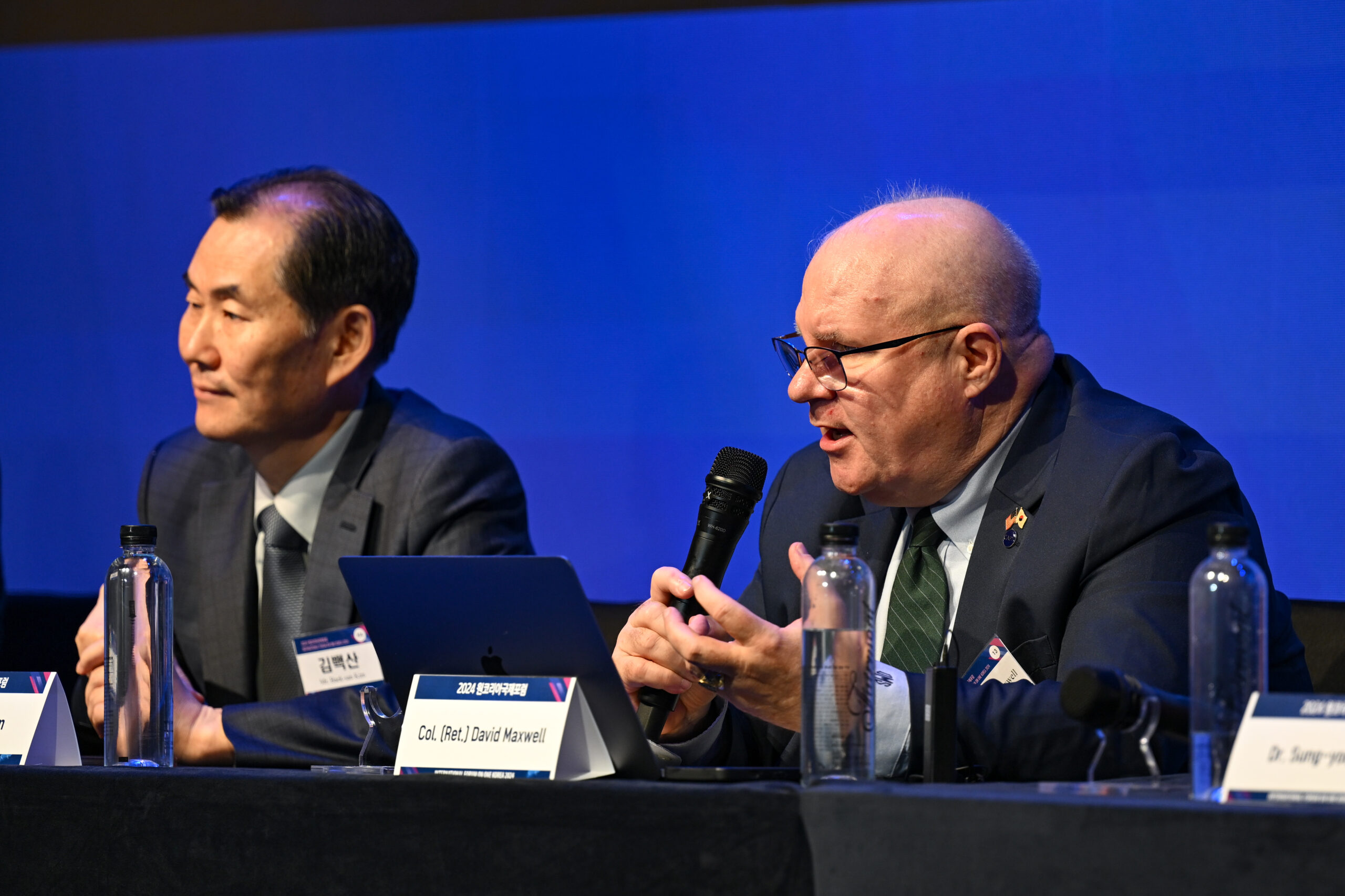 Two men in suits sit at a table with microphones and laptops, speaking at an International Forum panel discussion, envisioning the future of One Korea in 2024.
