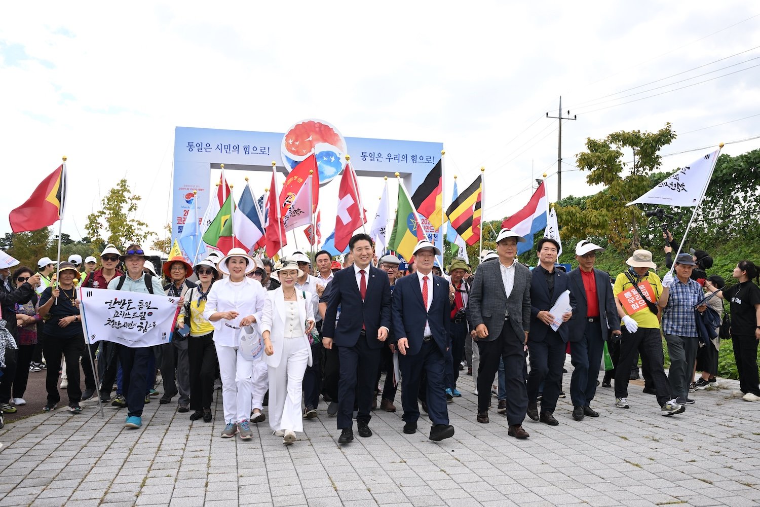 A diverse group of people, embodying the Korean Dream, marches down a path holding various national flags. Some wear suits, others casual clothing, under an archway with text and decorative elements.