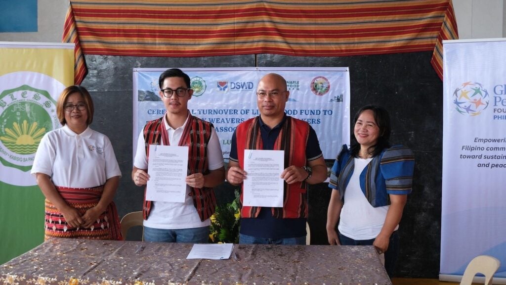 A group of five people stands together holding documents in front of a table, with banners highlighting the Livelihood Grant awarded to the Chumma-ay Weavers in the background.