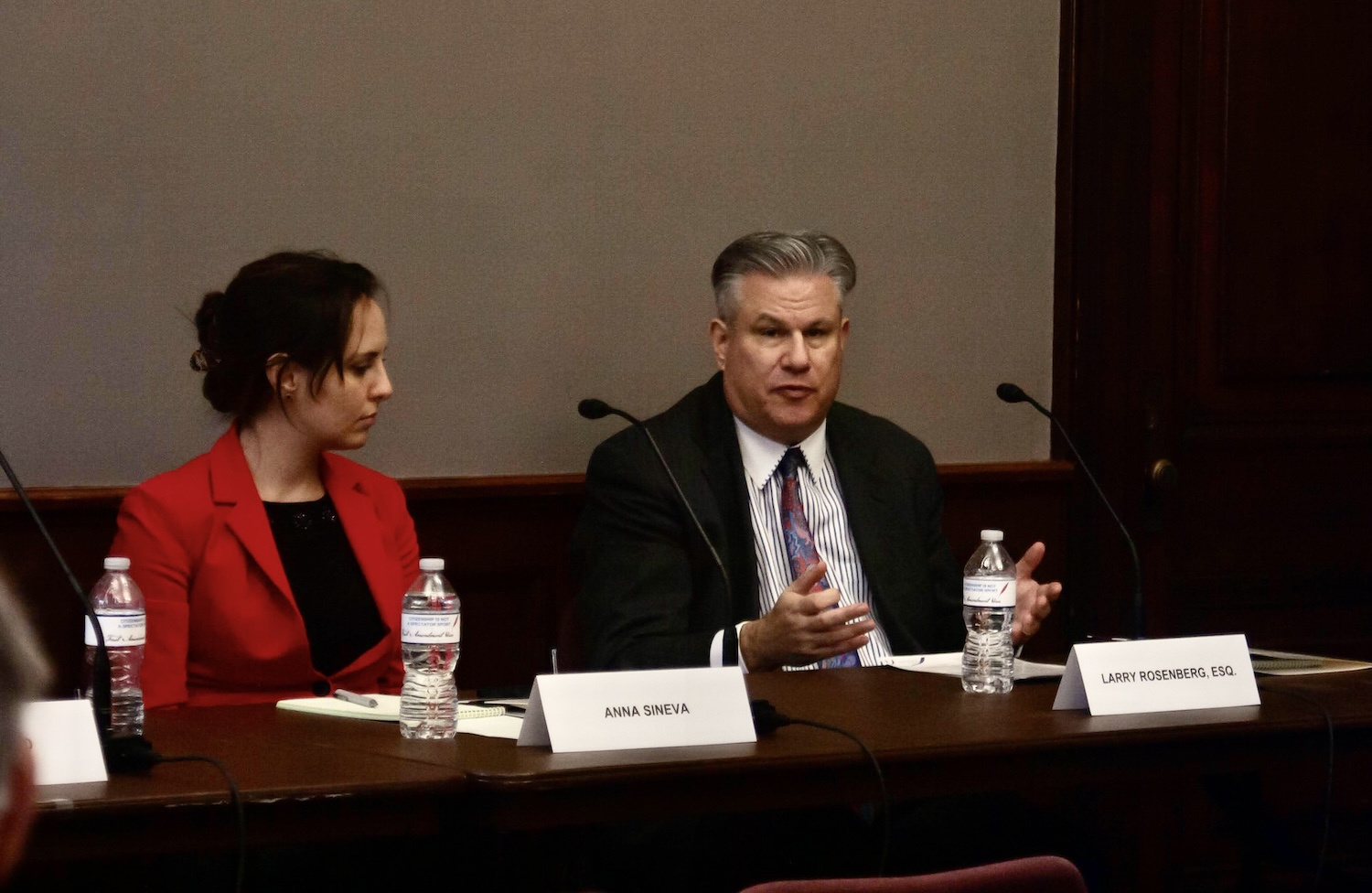 Anna Sneva and Larry Kossberg, Esq., are seated at a Capitol Hill conference table, engaging in a discussion on religious freedom. Nameplates in front of their microphones and water bottles underscore the importance of the dialogue taking place.