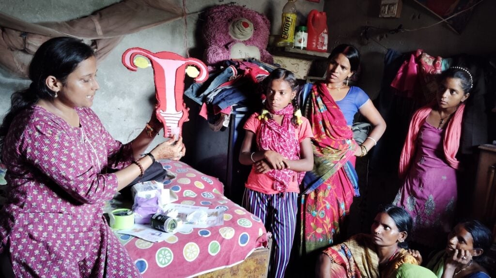 In the dimly lit room, a woman holds a uterus model, passionately explaining menstrual health to a group of women and girls from an urban slum.