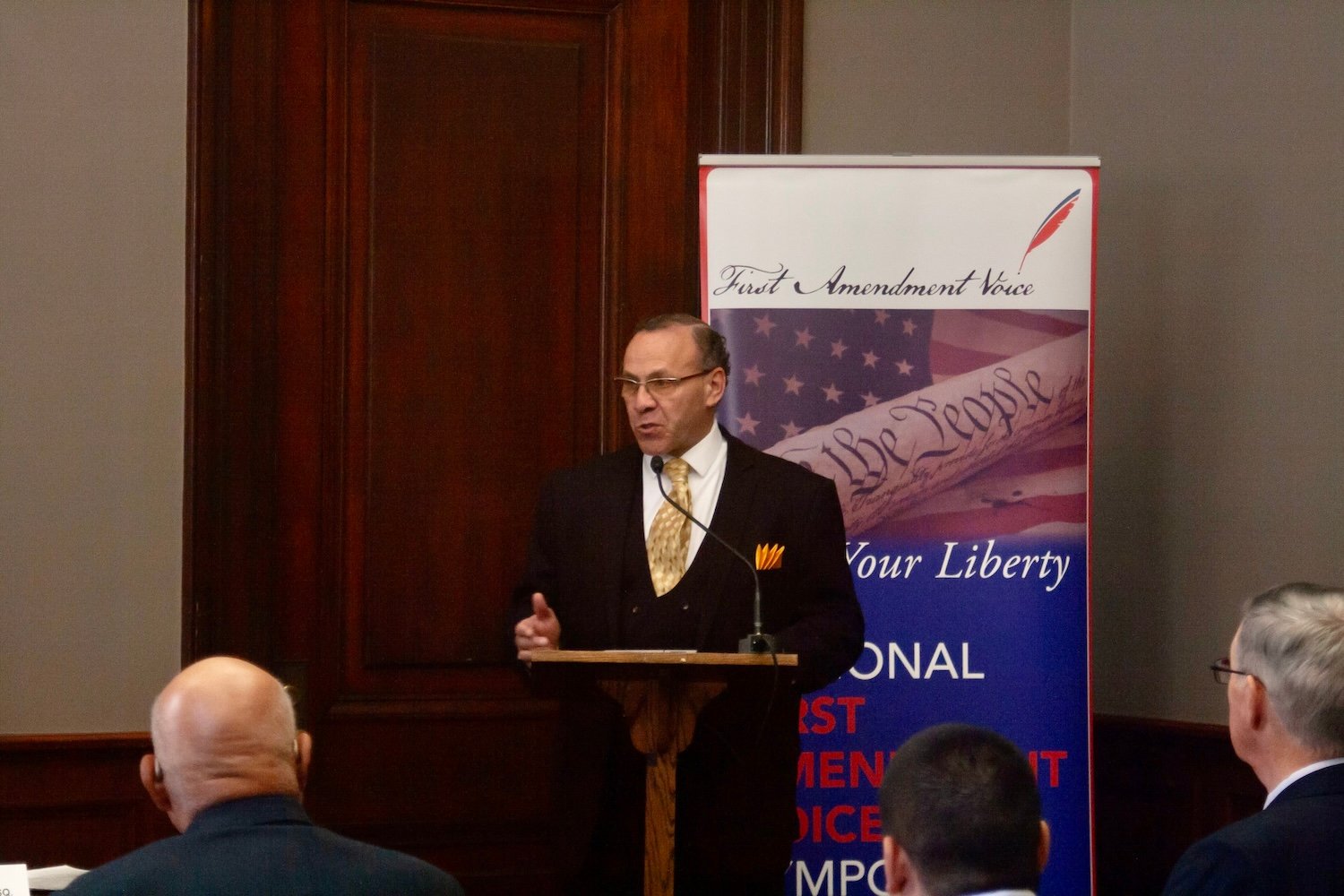 A man in a suit speaks at a podium in front of a "First Amendment Voice" banner, passionately addressing the importance of Free Speech. Several people are seated in the foreground, attentively listening to his words echoing through Capitol Hill.