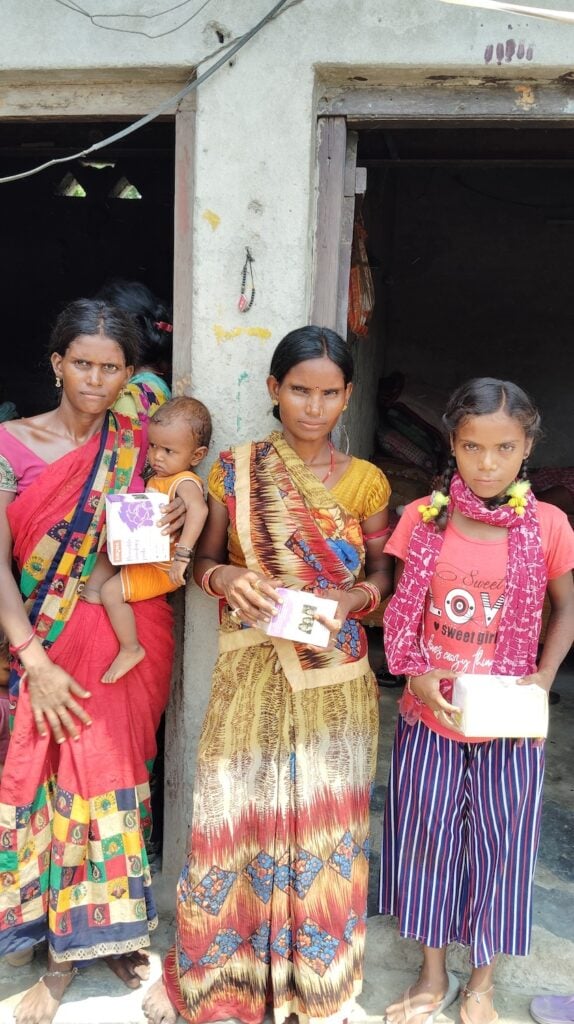 Three women and a child stand in front of a building, holding packaged items. In the vibrant scene, two women wear colorful saris, while the girl sports a T-shirt and skirt. Their gathering highlights GPF India's commitment to addressing menstrual health challenges in urban slums.