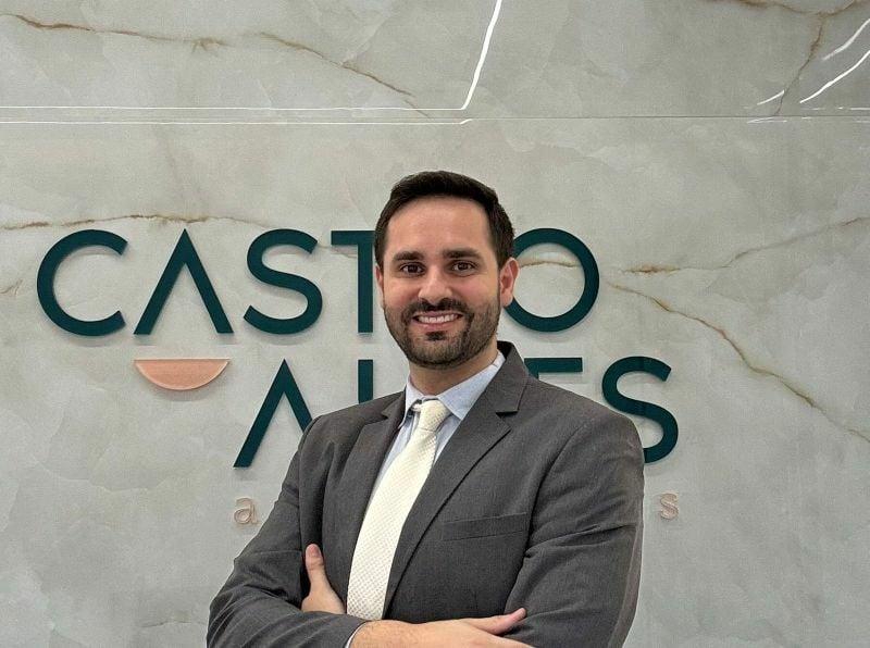 A man in a suit and tie stands with arms crossed in front of a sign that reads