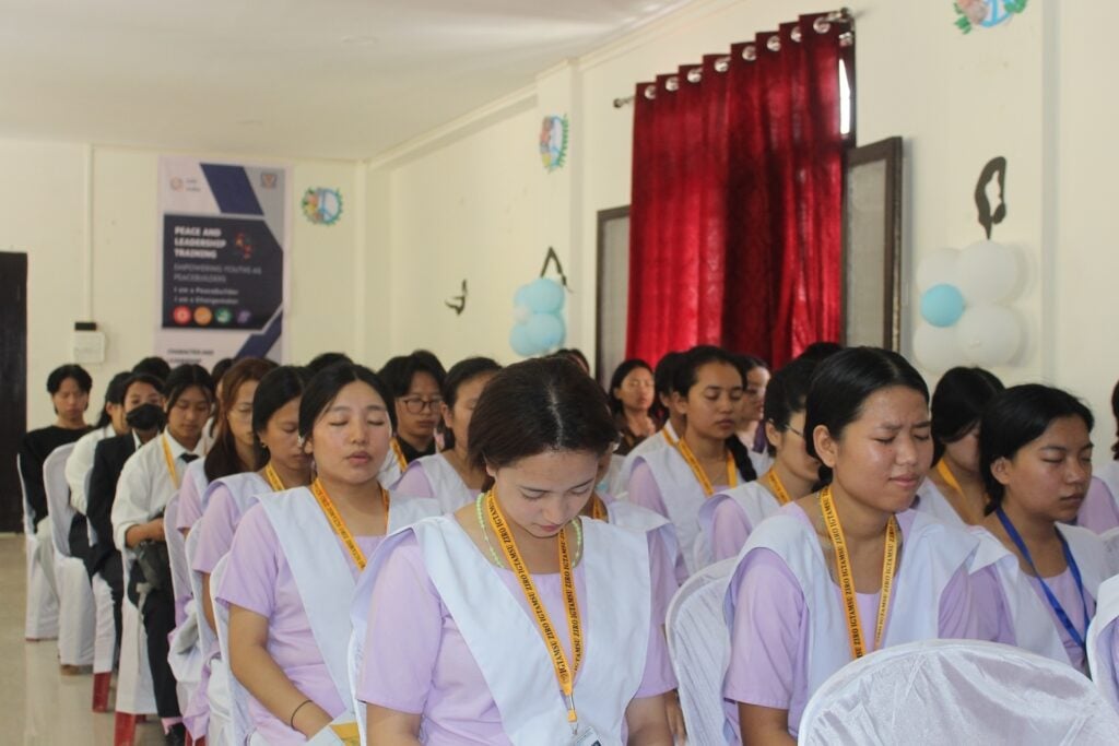 People in uniform sit in rows, some with eyes closed, in a room decorated with balloons and a banner on the wall celebrating peace and growth.