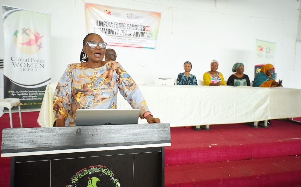 A woman stands speaking at a podium, while five women from the Families for Peace initiative sit at a table on stage, with a banner in the background. This empowering event takes place in Kaduna State.