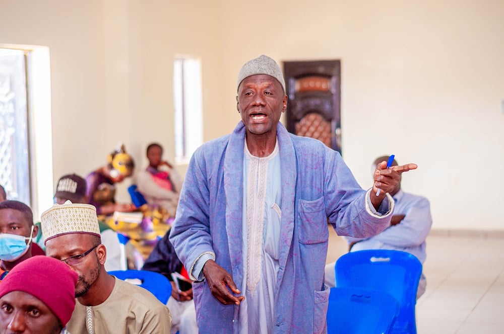 An elderly man in a robe speaks passionately about equitable access to education while standing among seated individuals in a room with blue chairs. Others listen, some wearing masks, as he highlights the importance of supporting vulnerable girls in Kaduna State.