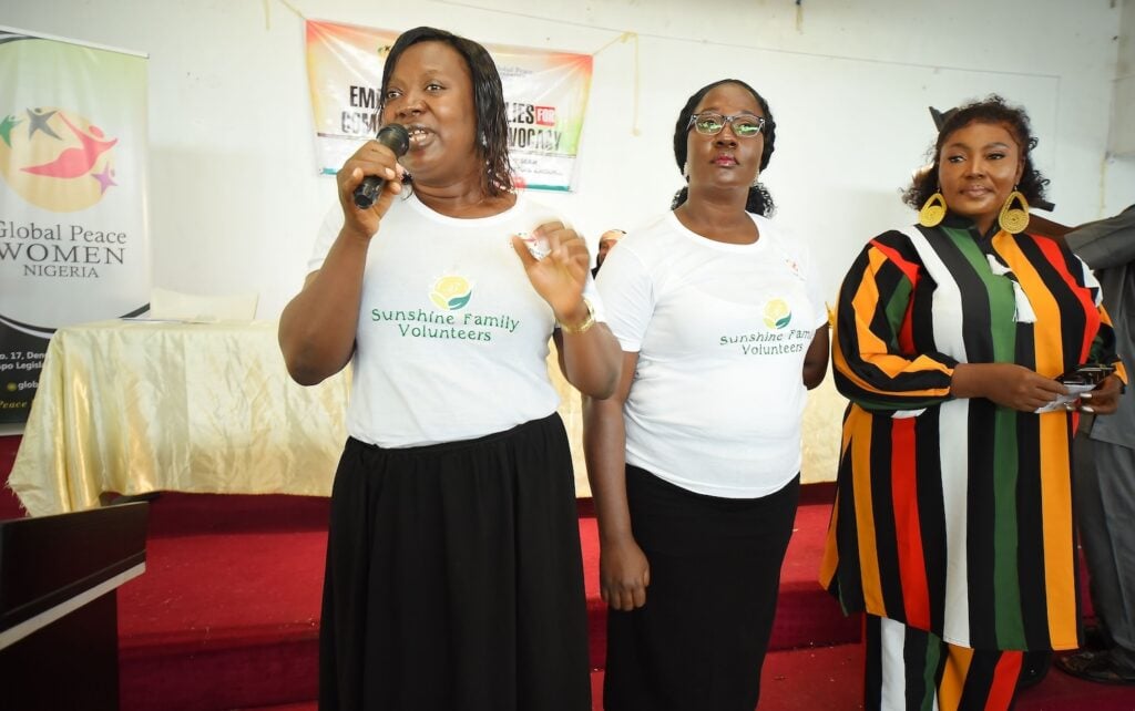 Three women stand on a stage; two in Sunshine Family Volunteers shirts, one holding a microphone. The third woman, wearing a colorful dress, embodies the spirit of Empowering Women. They are in front of banners promoting peace initiatives in Kaduna State.