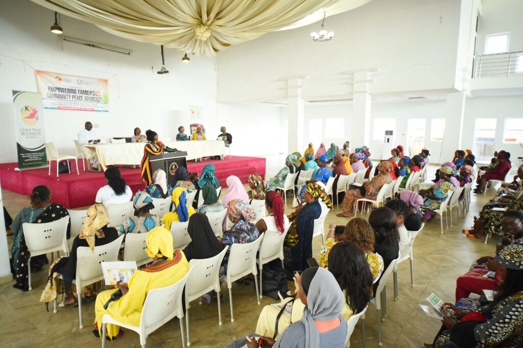 A group of people sits in rows facing a panel of speakers, heralded as Agents of Change, on a stage in a large room with a high ceiling. The event, held in Kaduna State, focuses on empowering women and fostering discussions that inspire transformative action.