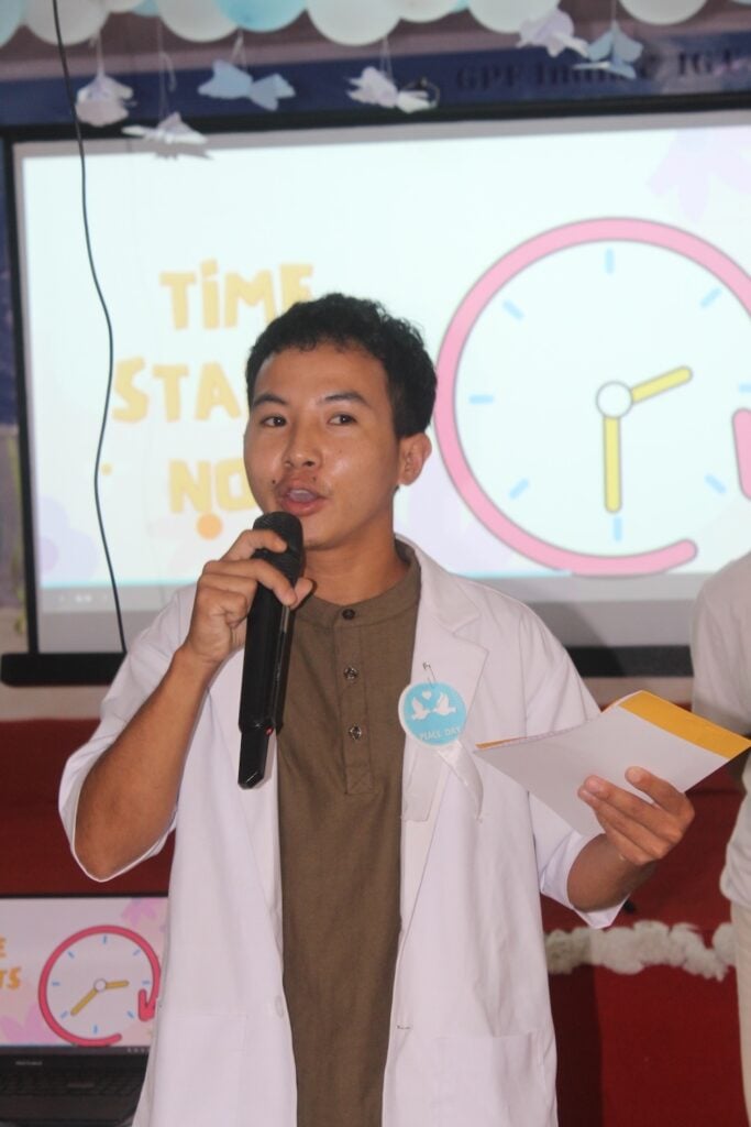 A person in a white coat speaks into a microphone, holding papers. A presentation screen in the background displays a clock and the phrase