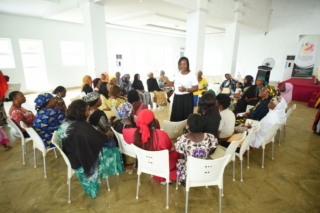 A woman stands speaking to a group seated in chairs during an empowering discussion in a large, well-lit room, inspiring families for peace in Kaduna State.