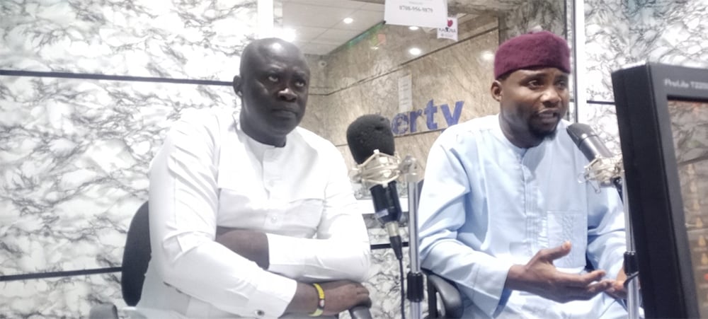 Two men, engaged in a spirited radio campaign, sit in a studio speaking into microphones. One is dressed in white, while the other wears light blue with a dark cap, discussing peaceful coexistence and harmony in Nigeria.