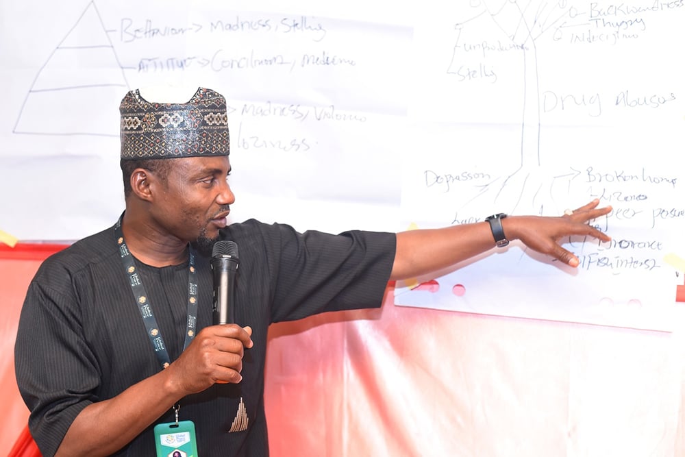 A man in traditional attire speaks into a microphone, pointing at a diagram on a board covered with handwritten notes, passionately discussing conflict analysis to empower the community.