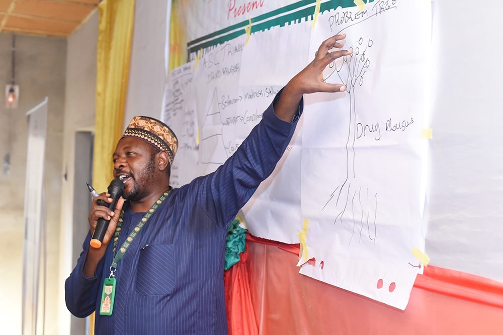 A man is speaking into a microphone, engaging the audience with a detailed conflict analysis as he points to a diagram on a large sheet of paper taped to a board in Hayin Banki.