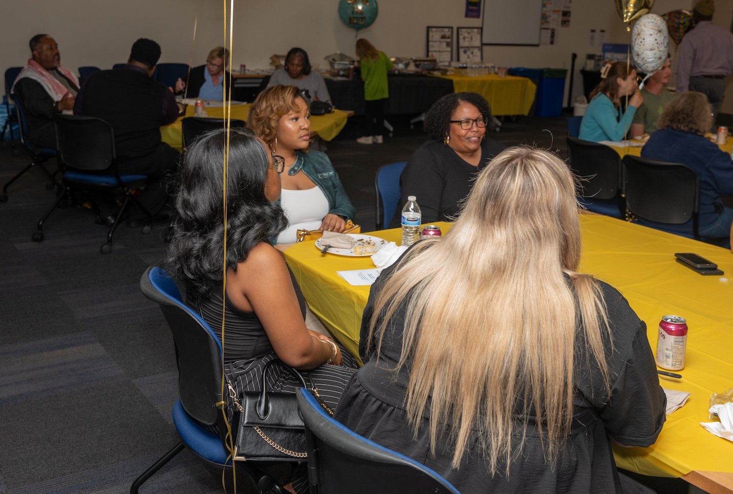 A group of Baltimore residents seated at tables covered with yellow tablecloths gathers in a room, attending a community engagement project focused on fostering positive race relations.
