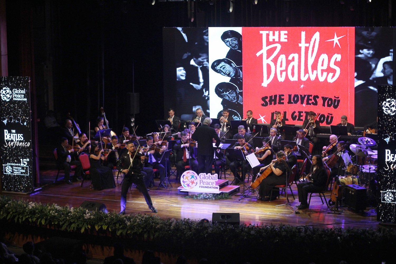 An orchestra performs on stage, led by a conductor and musicians dedicated to harmony. A large screen displays