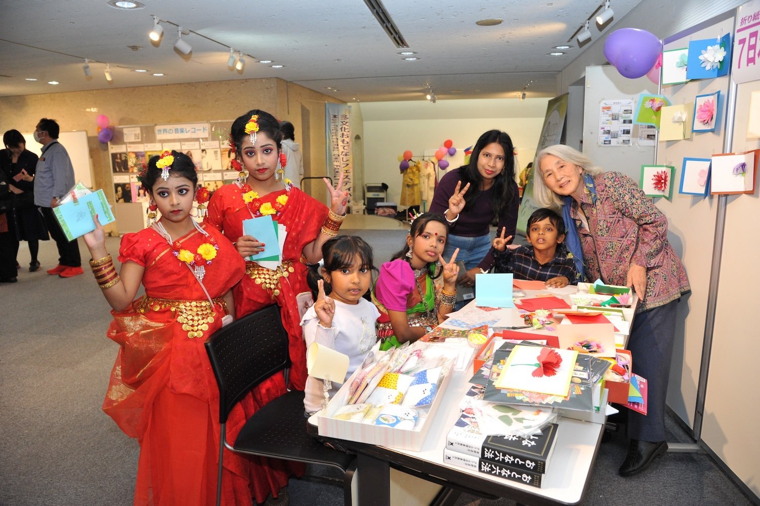 Children's Play A multicultural group, including children in colorful attire, gathers around a table with crafts and art supplies inside a room adorned with festive displays and balloons, celebrating the Family Festival 2024.