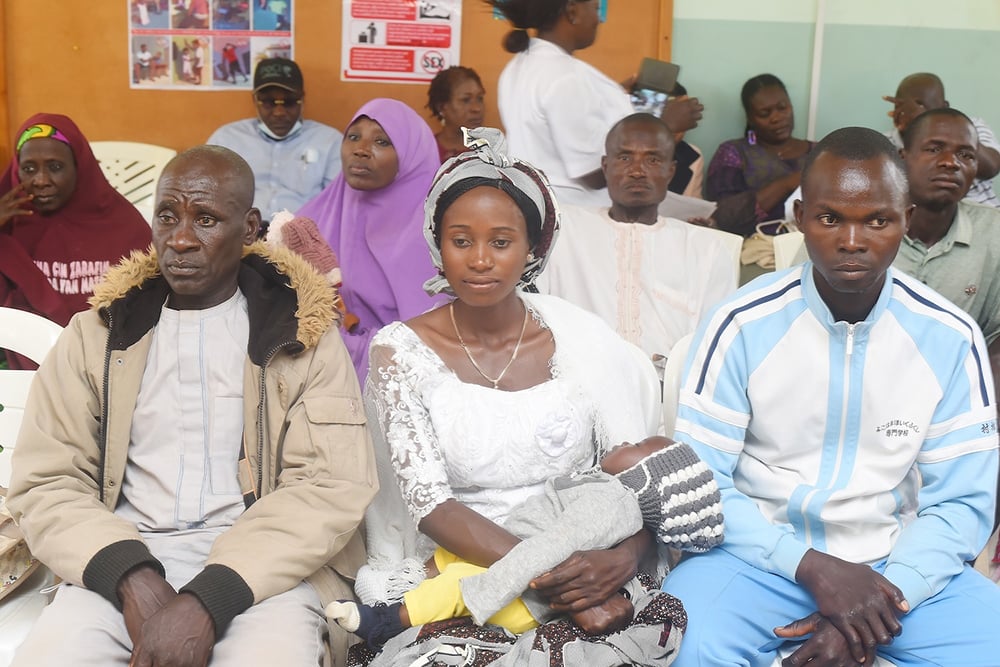 In a waiting area, a group sits together—a woman in a white dress cradles her baby, flanked by two men. Their shared presence symbolizes uniting communities against challenges faced in regions like Nigeria.